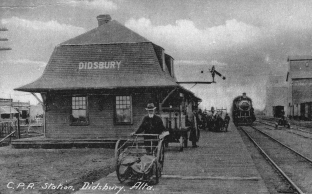Record ID PIC1567426769. 1924 Photo - There is a train by the station and a man pushing a mail cart. Grain elevators are on the east side of the tracks. Message on postcard reads Didsbury, Alta Dear Viola Well I spoke to grandma Shantz you can stay with her.